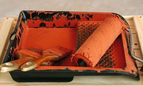 A paint tray with two paint brushes, and a paint roller in it, covered with orange paint.