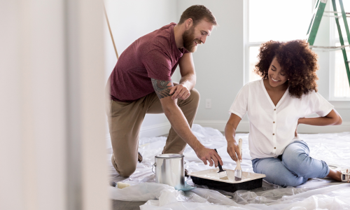 A couple sitting on the floor dipping their paint brushes in a paint tray preparing to paint the interior of a room.